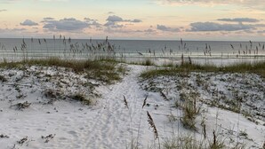 Un sentier bordant la plage, menant à l’océan, avec du sable et de l’herbe au premier plan.
