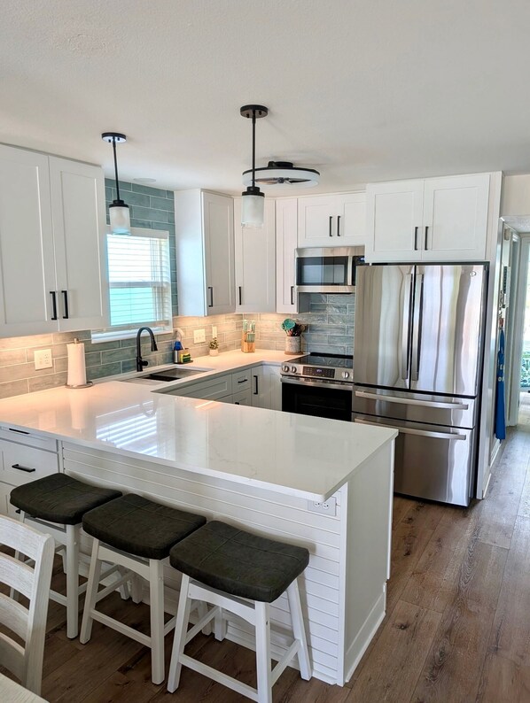 Une cuisine moderne avec des meubles blancs, un grand îlot central doté de tabourets, des appareils en acier inoxydable et un ventilateur de plafond.