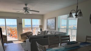 Un vaste espace de vie comprenant un canapé, une table à manger et des chaises, un ventilateur de plafond et de grandes fenêtres offrant une vue sur la plage.