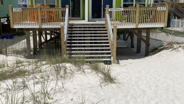 Une maison de plage à deux étages, à l’extérieur vert, portant les numéros 1239 et 1237, dotée d’une terrasse en bois avec des sièges d’extérieur et d’un parasol jaune.
