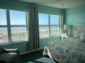 Una habitación con cama, una silla, una mesita de noche con lámpara y ventanas grandes con cortinas.