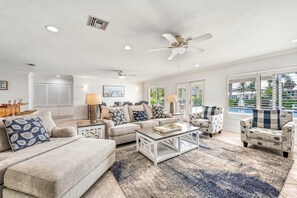 A spacious living room with a ceiling fan, a sofa set, armchairs, a coffee table, and a view of the outdoors through large windows.