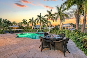 Zona de piscina con sillas de descanso y una mesa de comedor, rodeada de palmeras y vistas al atardecer.
