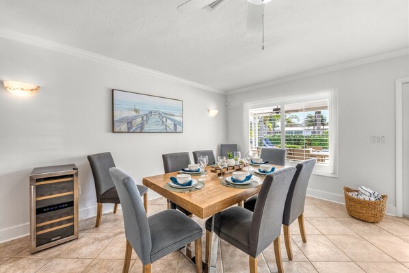 A dining area with a wooden table set for six, grey upholstered chairs, a painting of a beach, and a window with blinds.