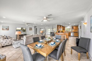 A spacious living area with a dining table set for four, a kitchen in the background, and a ceiling fan.