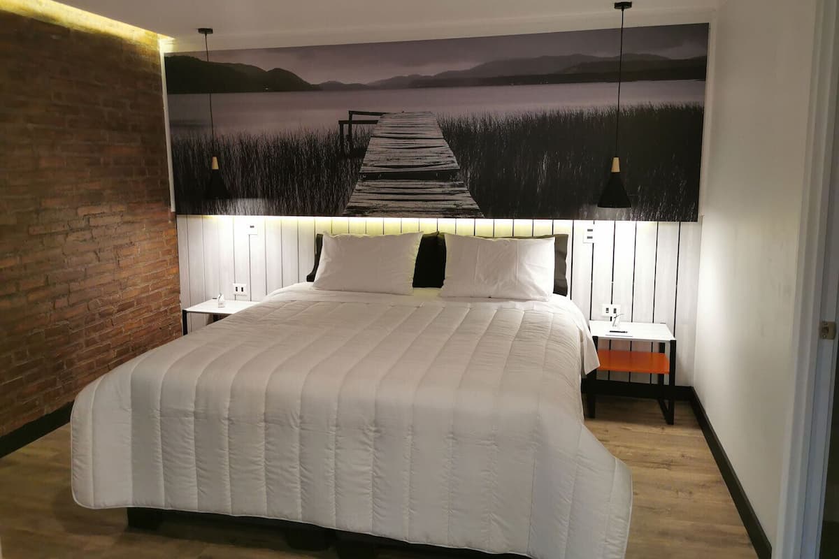 A bedroom with a large bed, a brick wall, a nightstand, a lamp, and a painting of a pier.