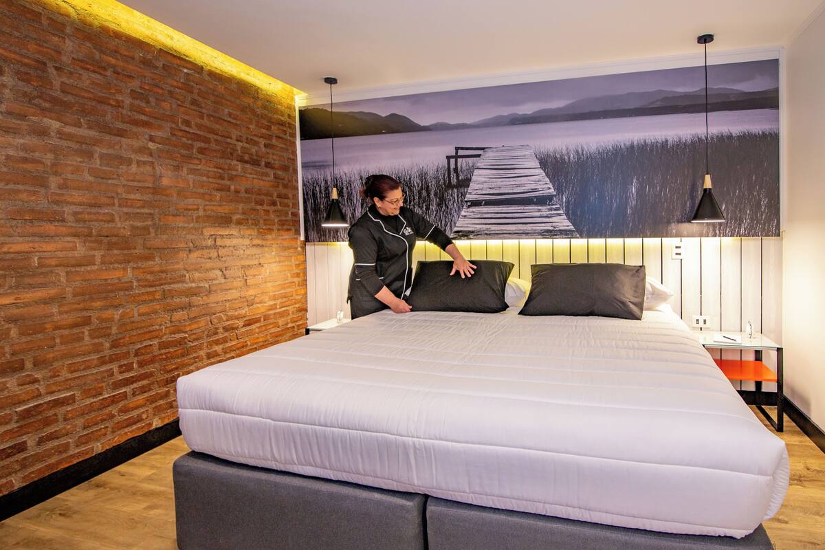 A modern hotel room with a large bed, a brick accent wall, and a scenic mural of a lake and pier.