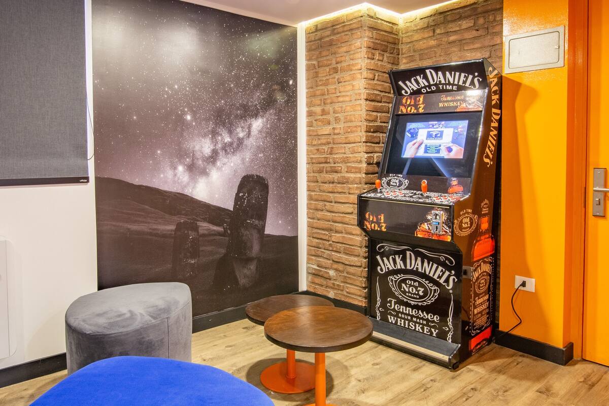 A room with a mural of a starry sky and a large stone head, a Jack Daniel's whiskey machine, a blue cushioned seating area, and wooden flooring.