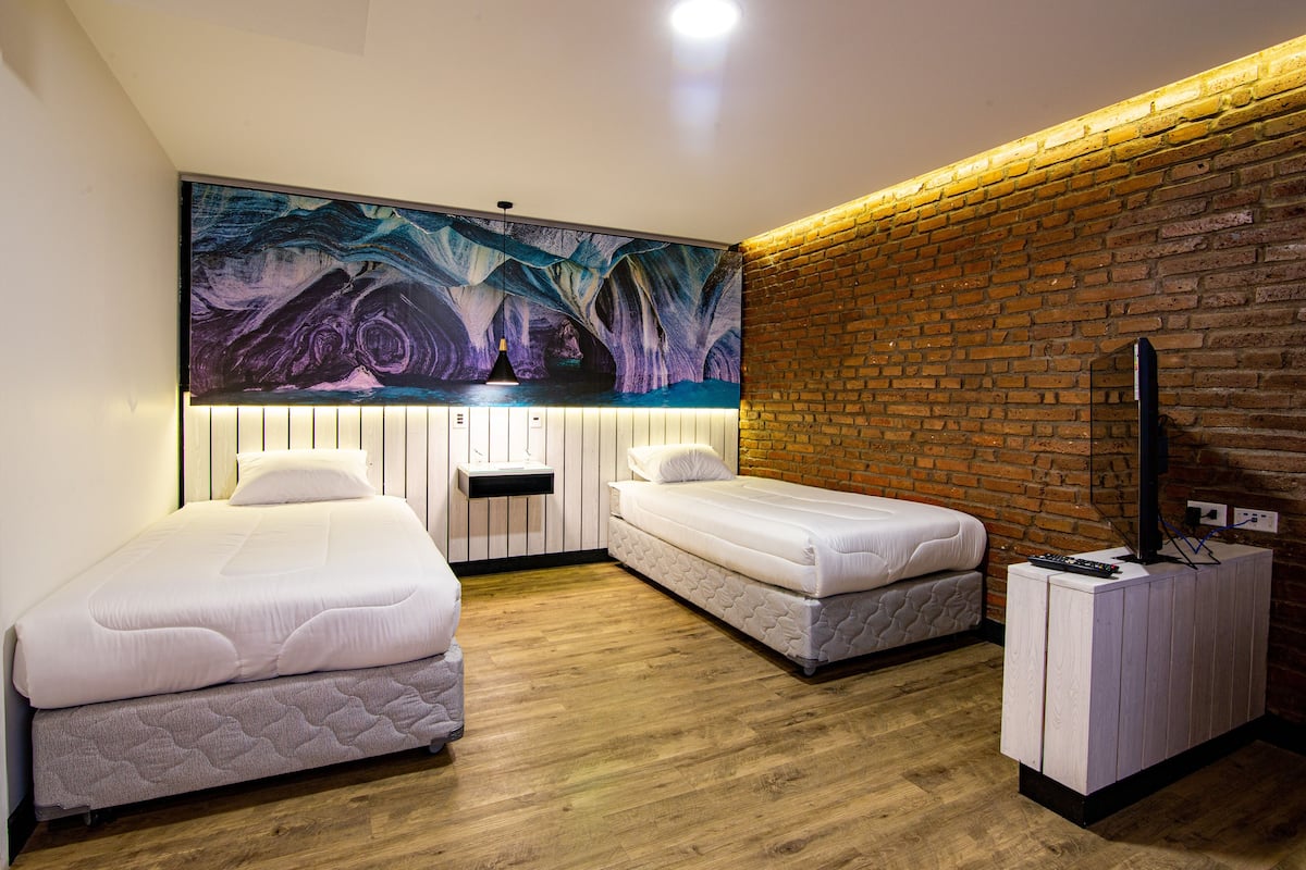A room with two beds, a large abstract painting, a brick wall, and a television on a wooden cabinet.