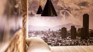 Une chambre avec une fresque représentant un paysage urbain sur le mur, un lit avec une literie blanche et deux lampes suspendues noires.