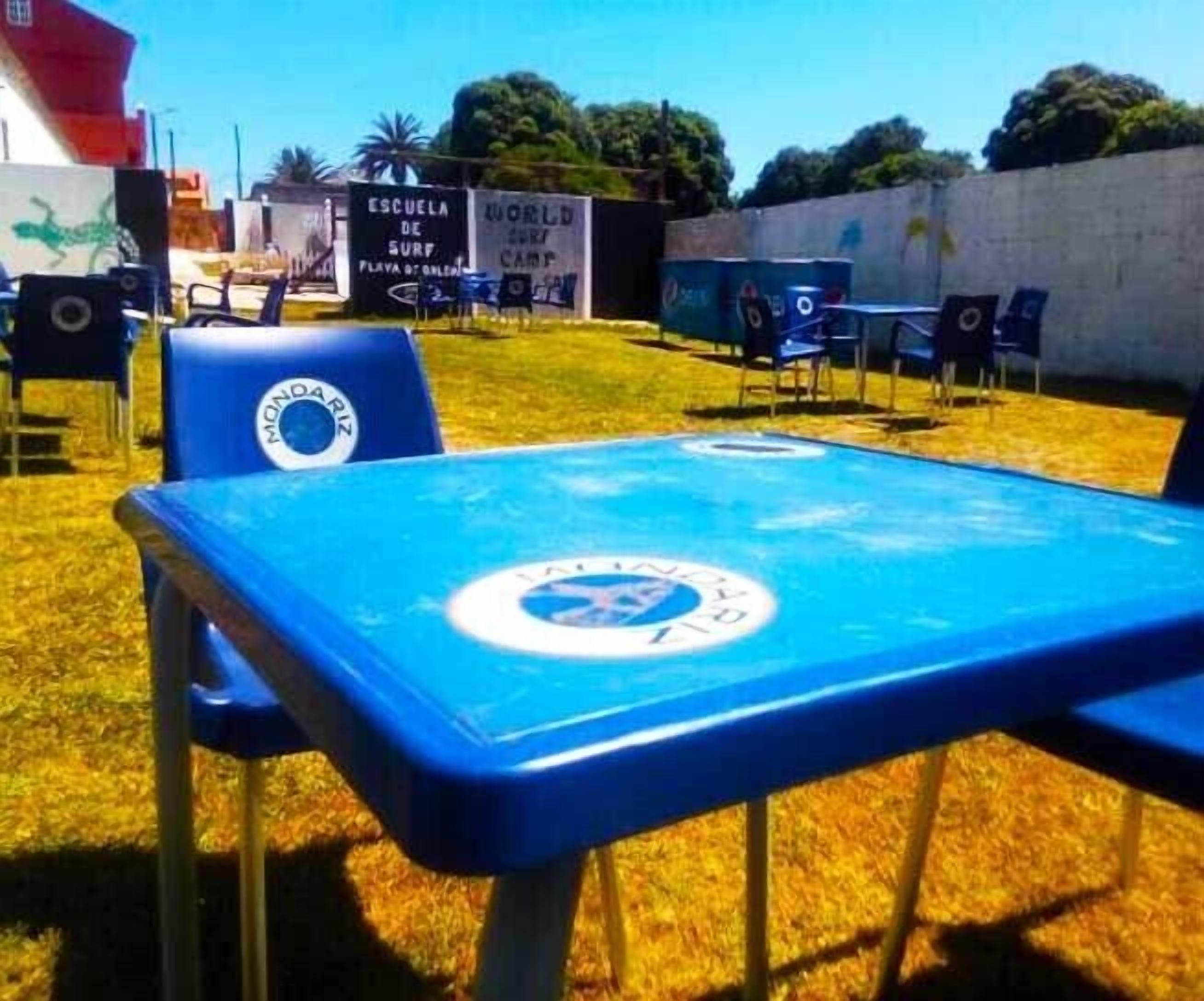 A blue table with a logo, surrounded by blue chairs, in an outdoor setting with a grassy area and a wall with graffiti.