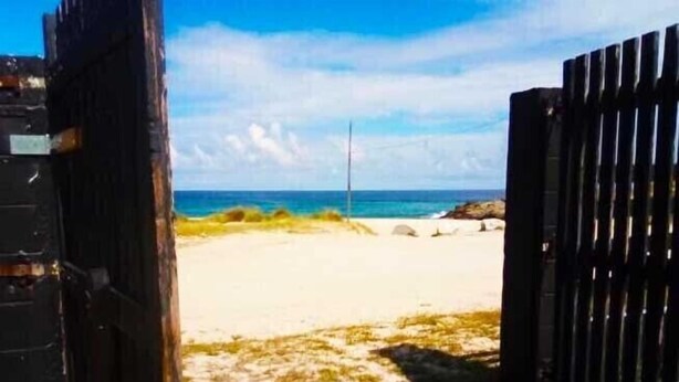 Blick auf einen Strand mit einem sandigen Pfad, der zum Meer führt, einem Holzzaun rechts und einem klaren Himmel.