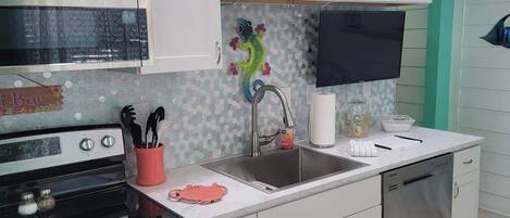 A modern kitchen with white cabinets, a stainless steel stove, a microwave, and a dishwasher. There is a sink with a faucet, a tiled backsplash, and a small television mounted above it.