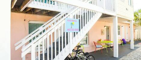 A multi-story building with a white staircase leading to a covered outdoor area. A sign advertises beach rentals with a phone number.