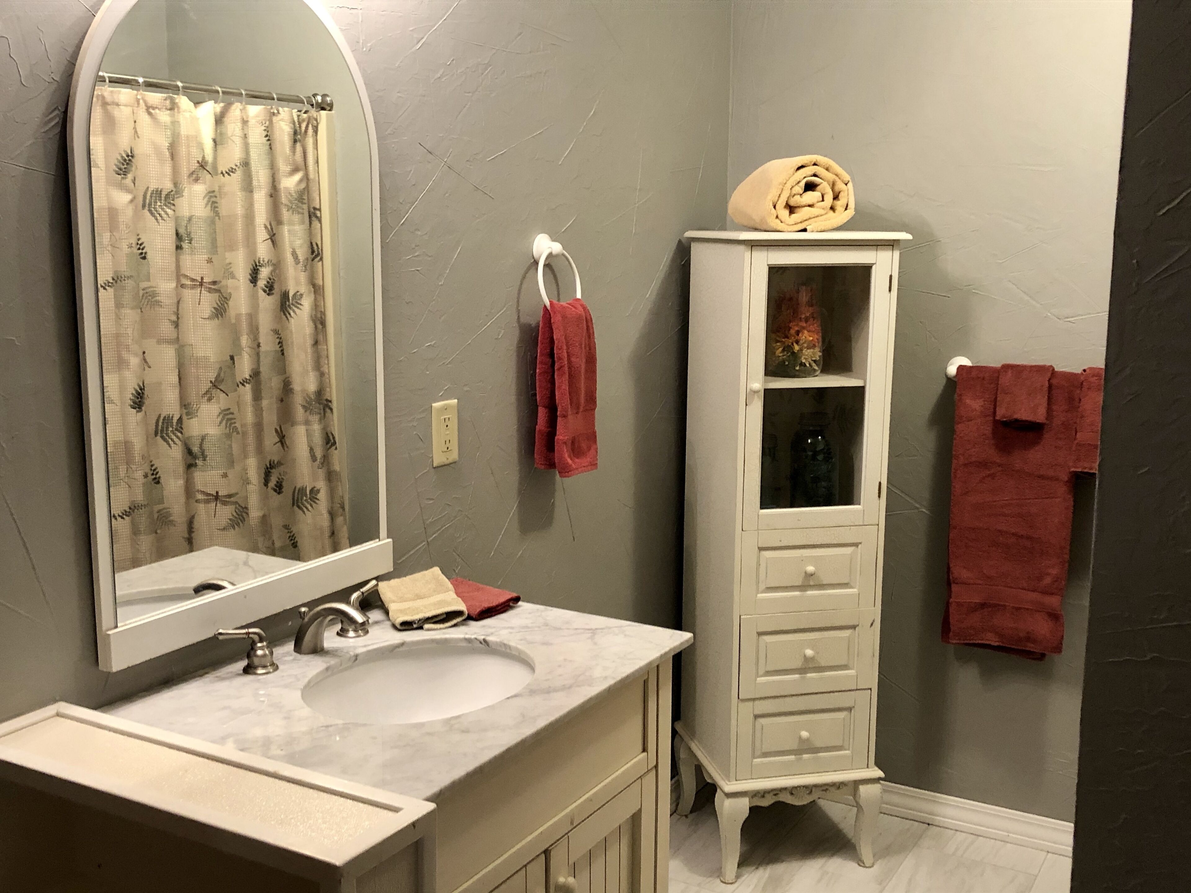 A bathroom with a marble sink, a white cabinet with glass doors, and a mirror with a leaf-patterned curtain.