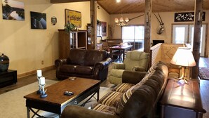 A cozy living room with a brown leather sofa, a green armchair, and a wooden coffee table. There is a dining area with a piano and a chandelier.