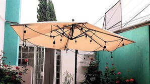 Pequena área de pátio ao ar livre com mesa e cadeiras sob um guarda-sol grande, rodeada por plantas em vasos e uma parede verde.