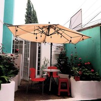 A small outdoor patio area with a table and chairs under a large umbrella, surrounded by potted plants and a green wall.