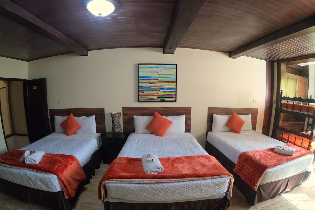 Quarto de hotel com três camas, cada uma com um cobertor vermelho e travesseiros brancos. Há um quadro na parede acima das camas.