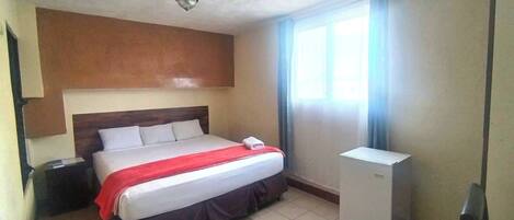 Une chambre d’hôtel avec un lit, une table de chevet, une commode et un luminaire au plafond.