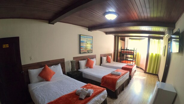 Quarto de hotel com três camas, cada uma com manta vermelha e almofadas brancas. Há um quadro na parede, uma luminária no teto e uma porta com um aviso.