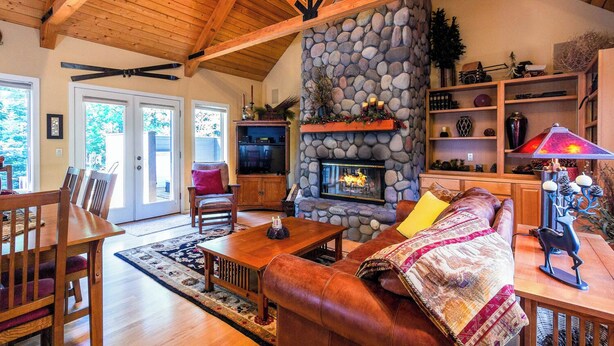 A cozy living room with a stone fireplace, wooden ceiling beams, and a dining area with a table and chairs.