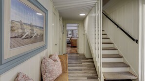 Um corredor com piso de madeira, escada com corrimão preto e um quadro com cadeiras de praia.