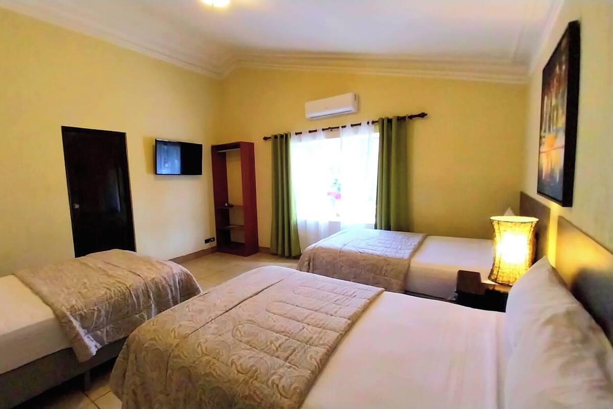 A hotel room with two beds, a wardrobe, an air conditioning unit, a television, and a window with curtains.