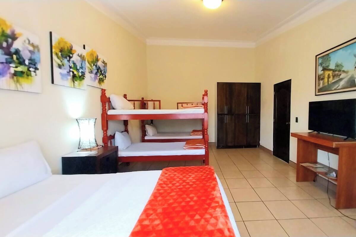 A hotel room with a large bed, a lower bunk bed, a TV, a wardrobe, and artwork on the walls.