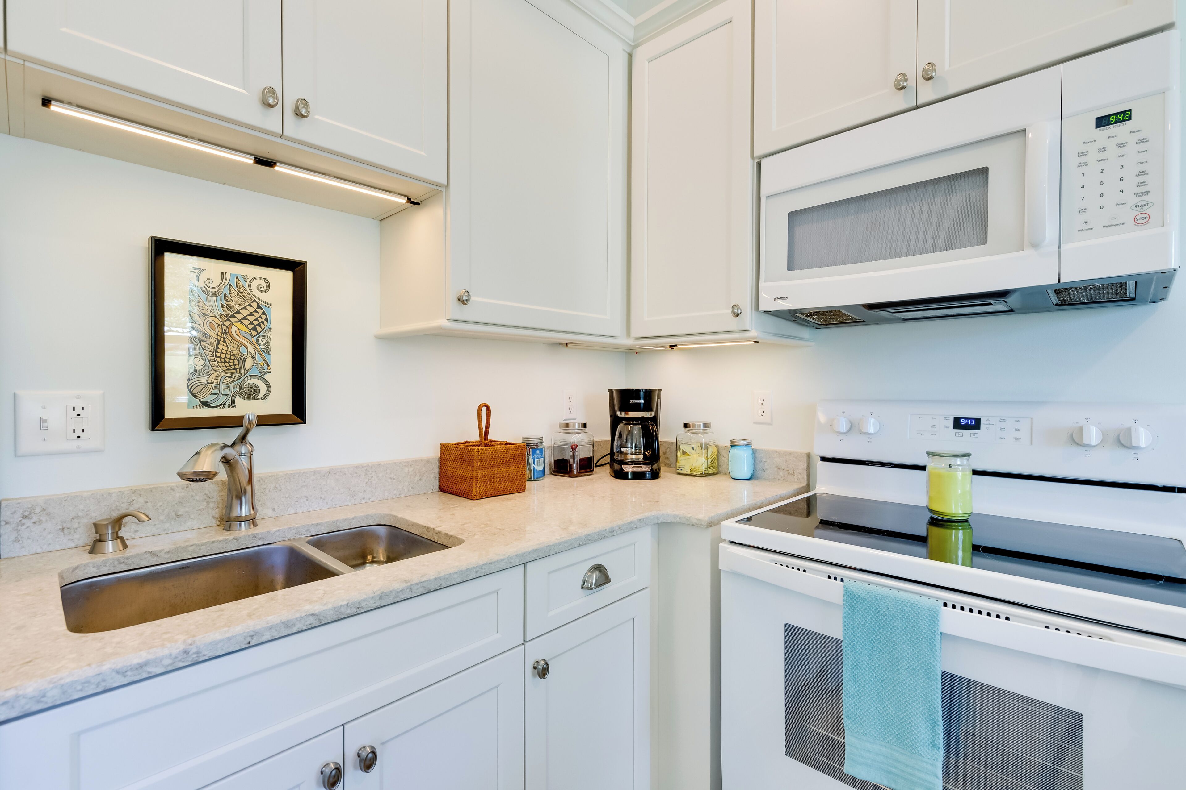 A kitchen with white cabinets, a stainless steel sink, a microwave, an oven, and a framed artwork on the wall.