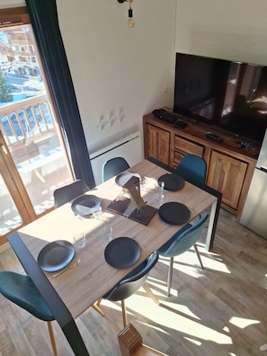 Ein Essbereich mit einem Holztisch, schwarzen Tellern und blauen Stühlen. Im Hintergrund befinden sich ein Fernseher und ein Holzschrank.