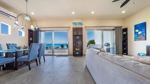 A spacious living area with a dining table and chairs, a sofa, a bookshelf, and a view of the ocean through large sliding doors.