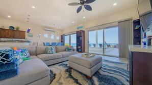 A spacious living area with a large sofa, an ottoman, and a dining table with chairs. There is a ceiling fan, a TV, and a view of the ocean through large sliding doors.