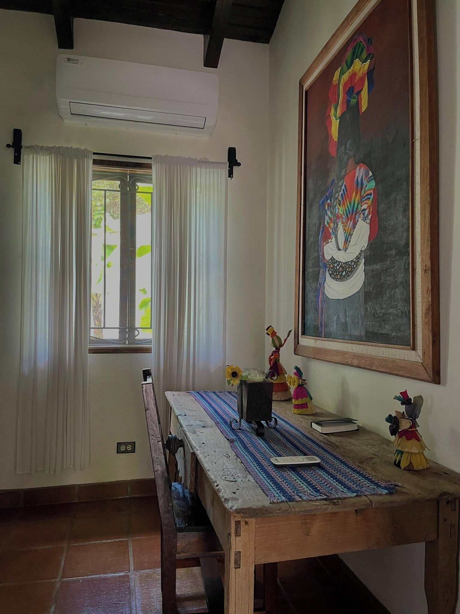 A room with a wooden table, a chair, a window with curtains, a painting on the wall, and a small statue on the table.