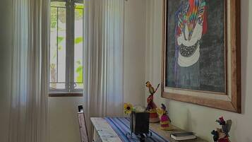 A room with a wooden table, a chair, a window with curtains, a painting on the wall, and a small statue on the table.