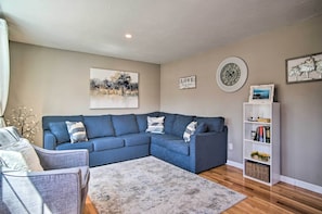 Een woonkamer met een hoekbank, een grijze fauteuil, een boekenkast en een schilderij aan de muur.