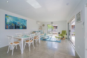 A spacious living area with a dining table set for six, surrounded by white chairs. A large abstract painting adorns the wall, and a green armchair is visible in the background. - Rose's Beach Pad - Matarangi Holiday Home (Matarangi)