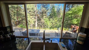 Une cuisine dotée d’une grande fenêtre donnant sur un patio aménagé avec des meubles d’extérieur et offrant une vue sur les arbres.