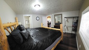 Un dormitorio con estructura de cama de madera, colchón negro, cabecera de madera, un reloj de pared, un espejo, un perchero y una ventana con persianas.