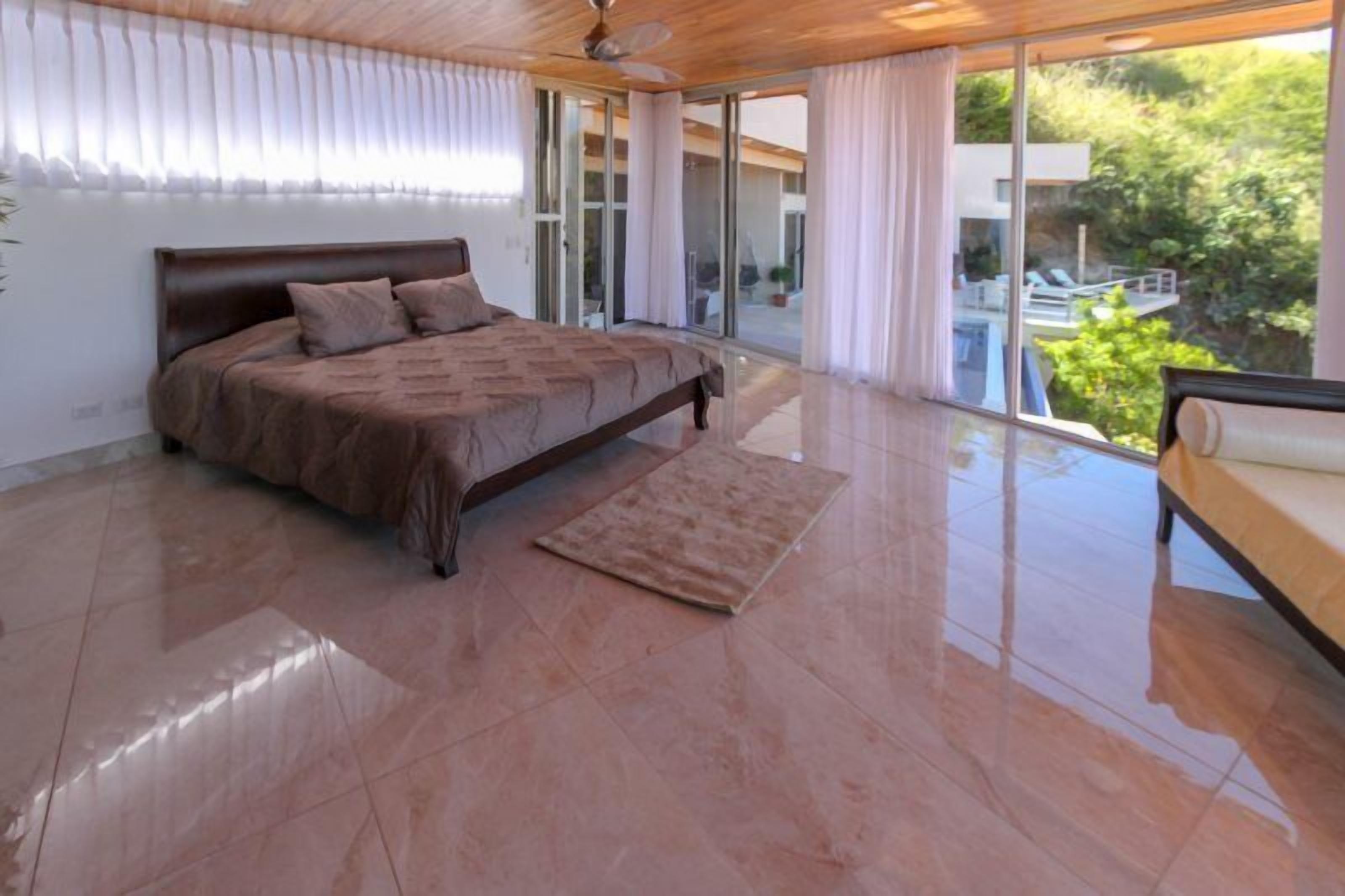 A spacious bedroom with a large bed, a wooden ceiling, and a view of a swimming pool and greenery outside.