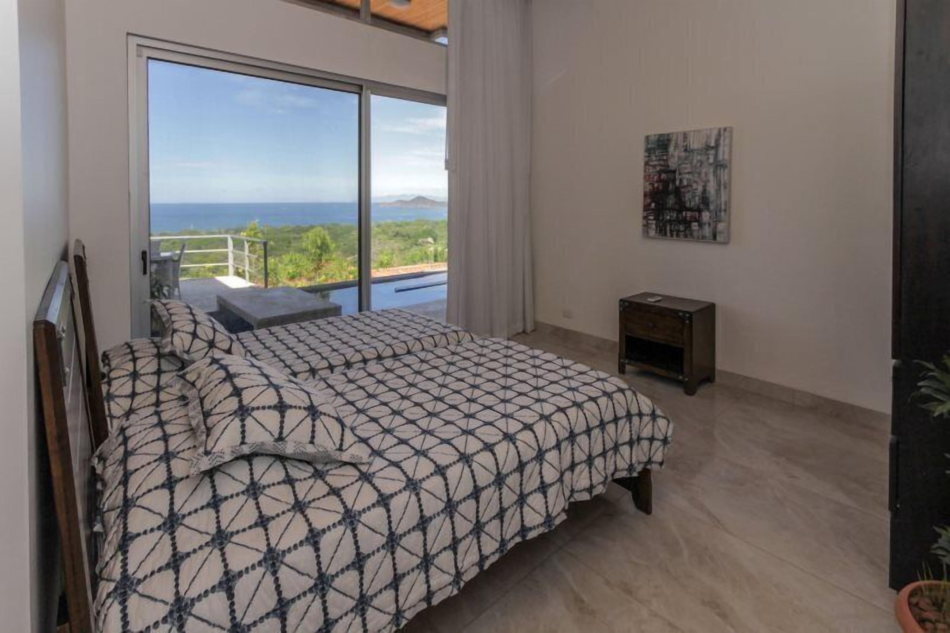 Une chambre à coucher avec un lit, une petite table, un tableau et une vue sur l’océan par une porte coulissante.