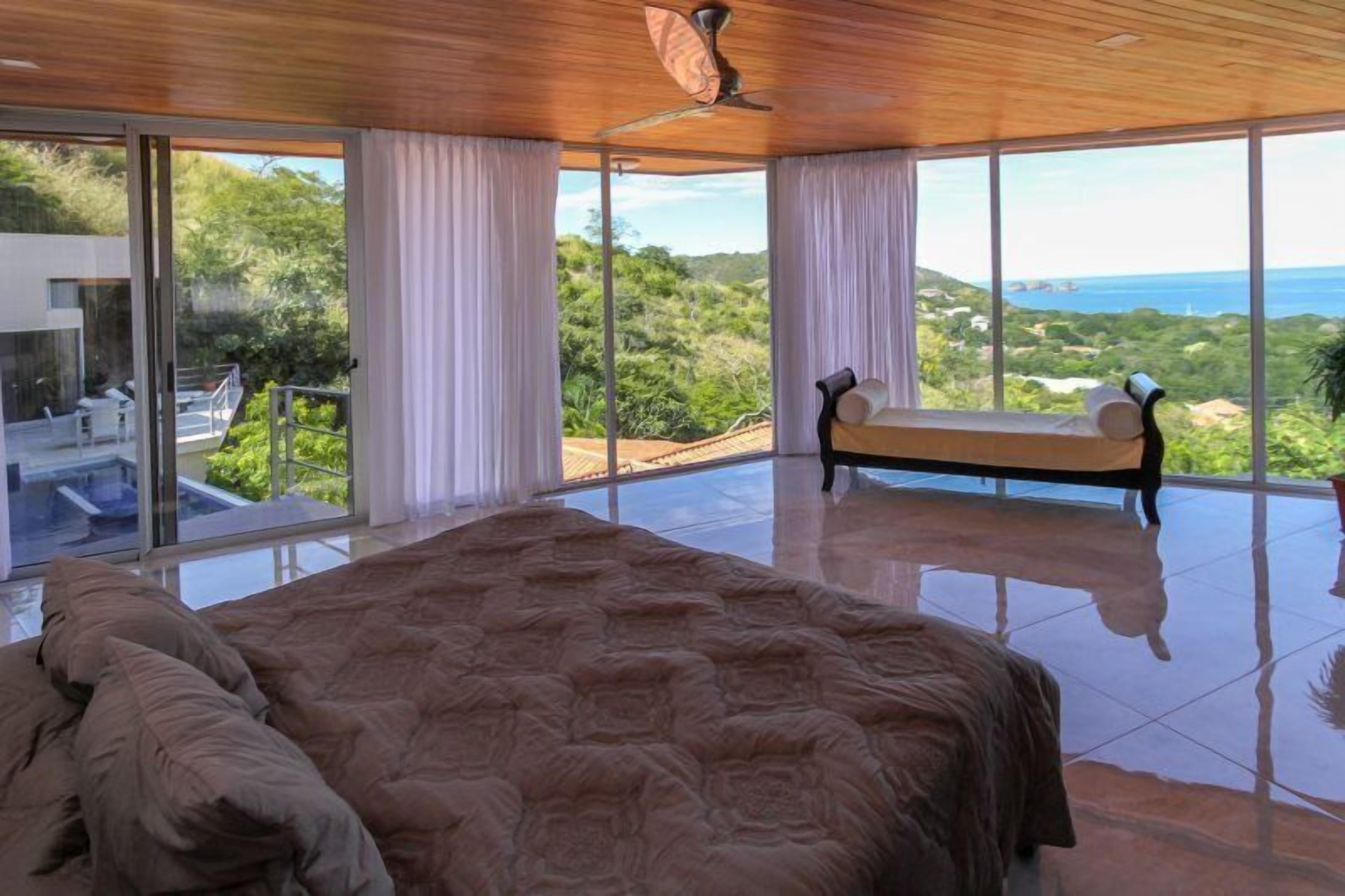 A spacious bedroom with a large bed, a bench, and a view of the ocean and greenery.
