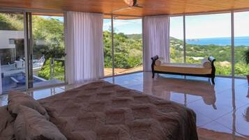 A spacious bedroom with a large bed, a bench, and a view of the ocean and greenery.