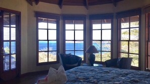 A bedroom with a large bed, a chair, a lamp, and a view of the ocean through large windows. - Casita Cole (Sardinal)