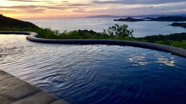 A swimming pool with a curved edge, surrounded by greenery and overlooking a body of water with distant landforms. - Casita Cole (Sardinal)