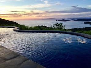 A swimming pool with a curved edge, surrounded by greenery and overlooking a body of water with distant landforms.