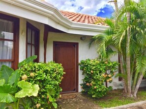 A house with a brown door, white walls, and a tiled roof. There are large green plants and a palm tree in the yard.