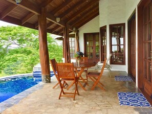 A covered outdoor area with a pool, wooden furniture, and a view of greenery. - Casita Cole (Sardinal)