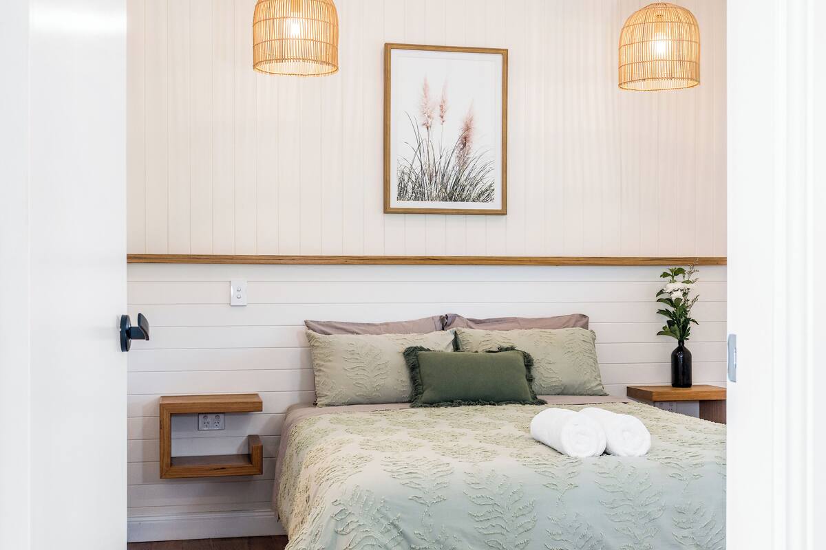 A neatly arranged bedroom with a bed, two bedside tables, a wall-mounted light fixture, and a framed picture above the bed.
