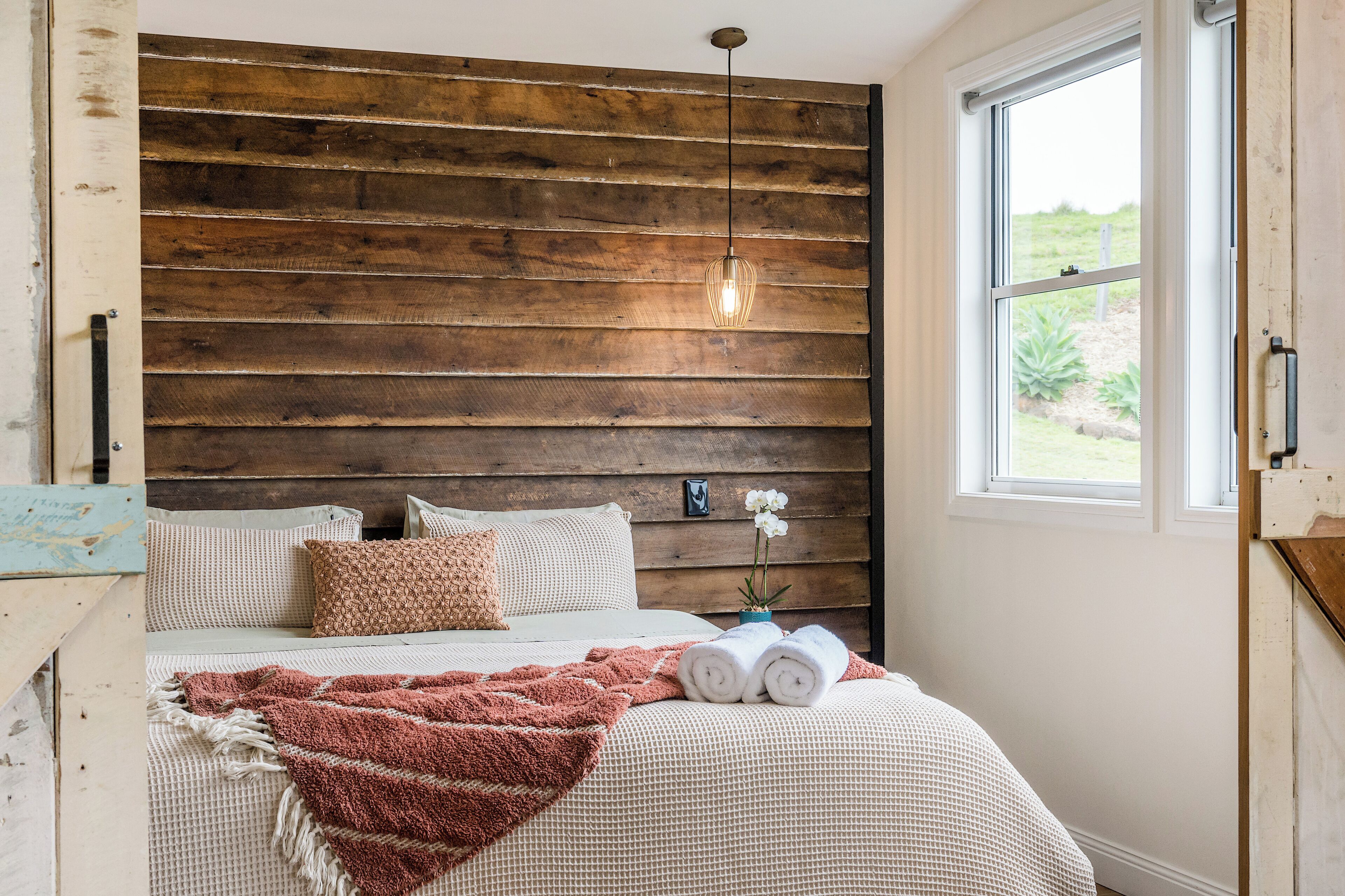A bedroom with a wooden headboard, a bed with white bedding, a red throw blanket, and a window with a view of greenery.